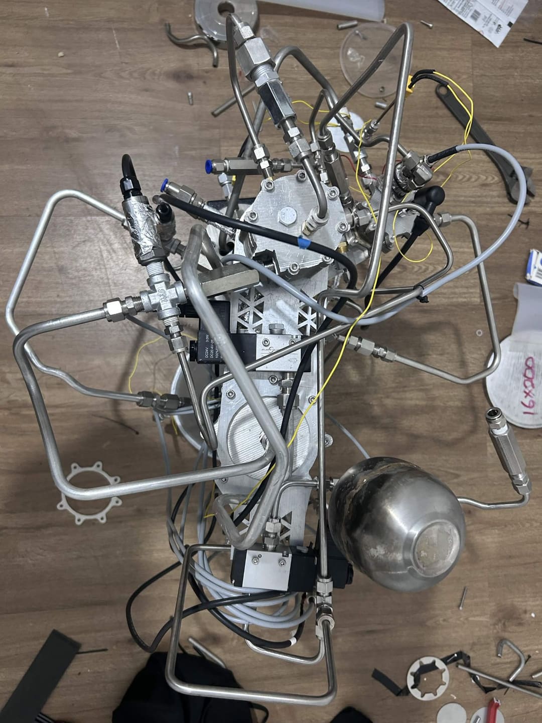 Top-down view of APEX propulsion development hardware on a wooden floor, with bent stainless tubing, fittings, cabling and a spherical vessel.