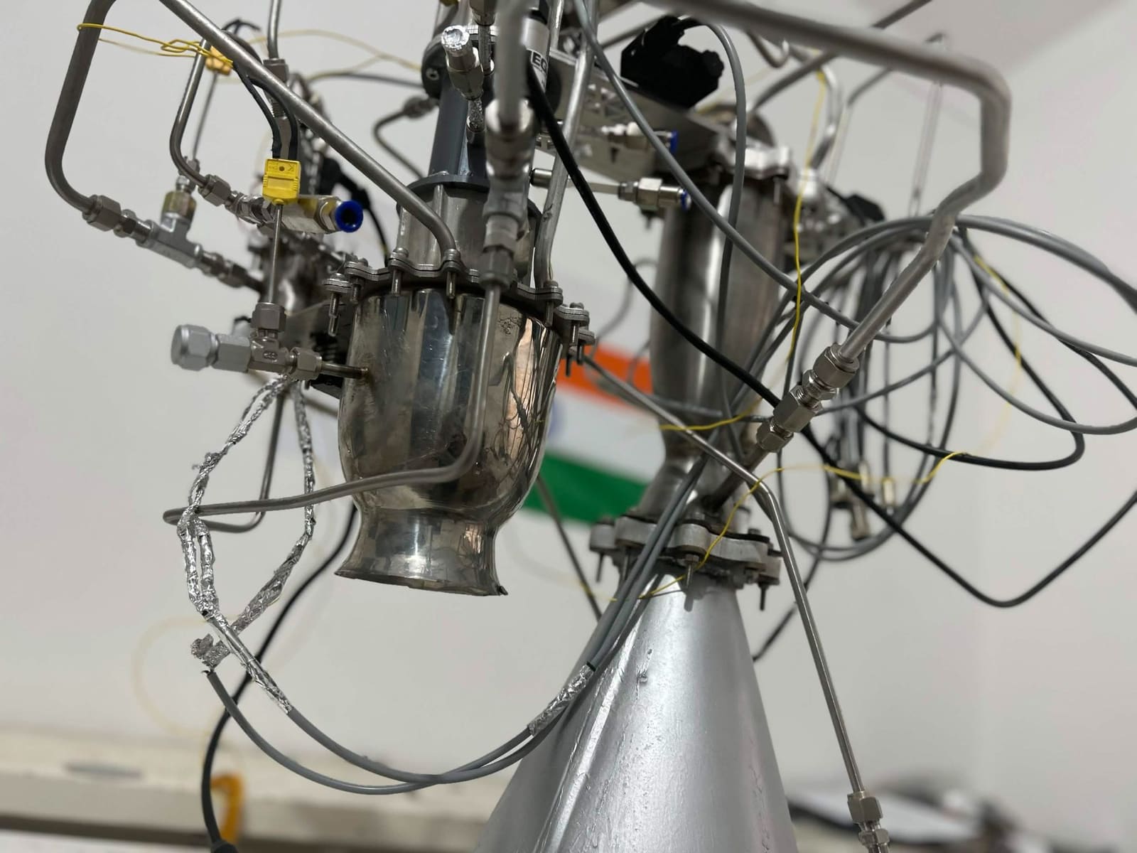 Low-angle view of APEX propulsion development hardware: polished metal sections, cabling and a large conical section, with the Indian flag in the background.